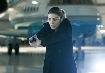 Mulher em casaco escuro, cabelo claro, aponta pistola com luz verde. No hangar, com avião ao fundo. Clima de ação e suspense.