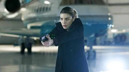 Lucifer – T01E13:Me leve de volta ao Inferno 1 Mulher em casaco escuro,cabelo claro,aponta pistola com luz verde. No hangar,com avião ao fundo. Clima de ação e suspense.