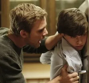 Um homem irritado segura e grita com um jovem, que está encolhido contra uma geladeira branca, parecendo assustado. Cena tensa.