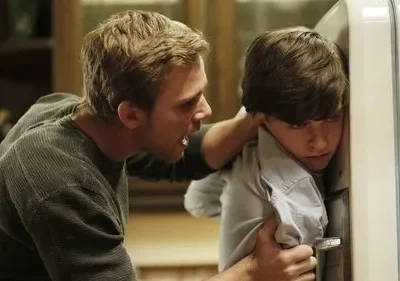 Um homem irritado segura e grita com um jovem, que está encolhido contra uma geladeira branca, parecendo assustado. Cena tensa.