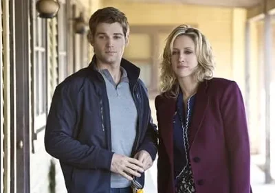 Homem de jaqueta azul e mulher loira de casaco roxo, ambos com expressões sérias, parados em uma varanda clara.