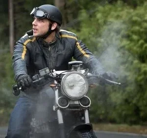 Homem com jaqueta de couro e capacete com óculos, em moto, olha preocupado. Fumaça paira, floresta verde ao fundo.