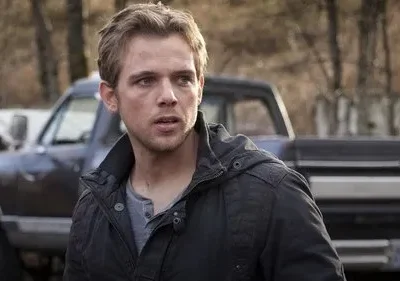 Jovem loiro de casaco escuro, com rosto sujo, olha tenso. Ao fundo, picapes velhas em cenário rural. Clima de suspense.