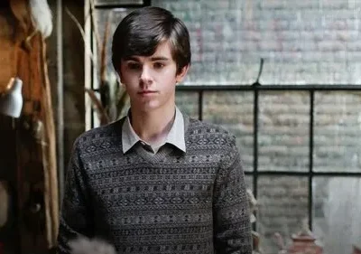 Jovem pálido de suéter estampado e expressão séria em quarto rústico, com janela grande ao fundo. Clima sombrio.