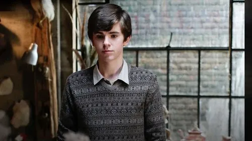 Motel Bates – T01E08:Um garoto e seu cão 1 Jovem pálido de suéter estampado e expressão séria em quarto rústico,com janela grande ao fundo. Clima sombrio.