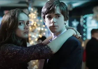 Jovem casal em festa com luzes. A garota, com cânula nasal, abraça o rapaz. Ambos têm expressões intensas, quase melancólicas.