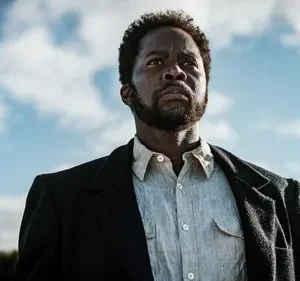 Um homem negro sério com barba e casaco, olhando para o céu azul. Fundo de campo aberto, clima determinado e pensativo.