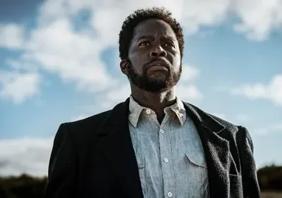 Um homem negro sério com barba e casaco, olhando para o céu azul. Fundo de campo aberto, clima determinado e pensativo.