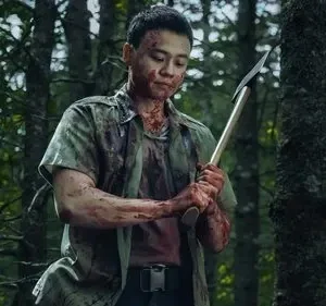 Homem com o rosto e roupas ensanguentados, segurando um machado, parado em uma floresta escura. O clima é tenso e perigoso.