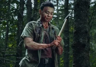Homem com o rosto e roupas ensanguentados, segurando um machado, parado em uma floresta escura. O clima é tenso e perigoso.