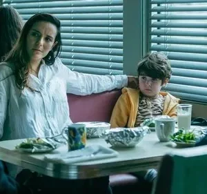 Mulher de camisa branca e menino sério em lanchonete, com dois homens em frente. Luz baixa, atmosfera sombria.