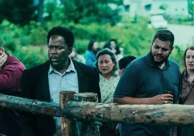 Um grupo diverso de pessoas observa algo com seriedade e apreensão, apoiados numa cerca de madeira em ambiente rural.