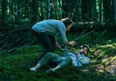Dois homens numa floresta musgosa e escura. Um deitado no chão, assustado, enquanto o outro o segura. Atmosfera sombria e dramática.