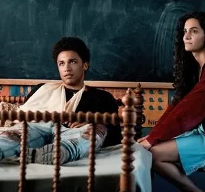 Um jovem deitado na cama e uma jovem sentada, olhando para lados opostos. Fundo com quadro-negro e cabeceira de madeira. Clima pensativo.