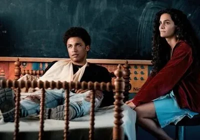 Um jovem deitado na cama e uma jovem sentada, olhando para lados opostos. Fundo com quadro-negro e cabeceira de madeira. Clima pensativo.