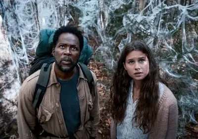 Homem e mulher assustados em floresta sombria, coberta por densas teias brancas. Clima de suspense/terror.
