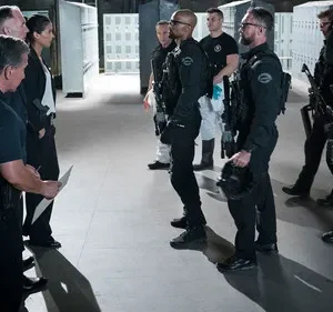 Equipe tática uniformizada em sala de treinamento. Um homem forte e uma mulher conversam, enquanto um grupo armado aguarda. Telas de segurança ao fundo.