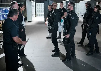 Equipe tática uniformizada em sala de treinamento. Um homem forte e uma mulher conversam, enquanto um grupo armado aguarda. Telas de segurança ao fundo.