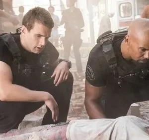 Dois homens em coletes táticos ajoelhados em escombros, examinando uma vítima. Cena de desastre, clima tenso.