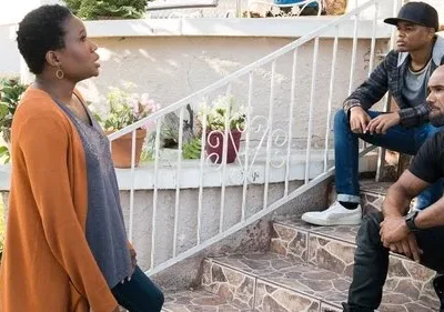 Mulher de laranja em pé; dois homens, um de boné, sentados em degraus externos com plantas. Expressões sérias sugerem diálogo tenso.