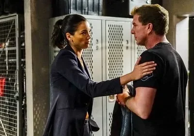 Uma mulher de blazer toca o ombro de um homem em camiseta preta. Em um vestiário, ela o conforta com uma expressão séria.