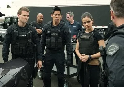 Quatro policiais LAPD SWAT, três homens e uma mulher, em uniformes táticos escuros, se reúnem ao ar livre com veículos.