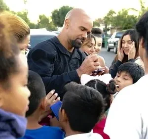 Homem careca barbudo em uniforme escuro assina autógrafos para crianças e jovens entusiasmados em uma rua residencial.