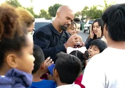 Homem careca barbudo em uniforme escuro assina autógrafos para crianças e jovens entusiasmados em uma rua residencial.