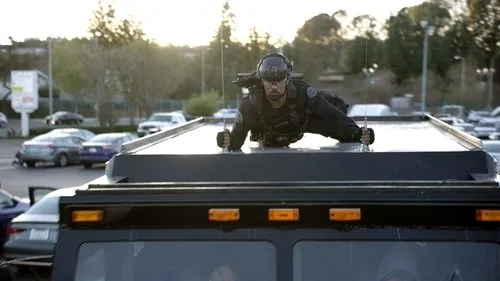 Homem de uniforme tático e capacete,deitado no teto de um veículo escuro em um estacionamento,ao pôr do sol. Clima tenso.