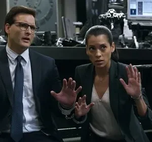 Um homem e uma mulher algemados com as mãos erguidas em um cenário sombrio e tecnológico. Ambos parecem assustados.