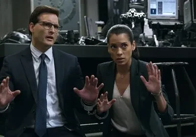 Um homem e uma mulher algemados com as mãos erguidas em um cenário sombrio e tecnológico. Ambos parecem assustados.