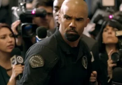 Um policial SWAT careca e barbudo, com expressão séria, em meio a uma multidão de jornalistas com câmeras e microfones.