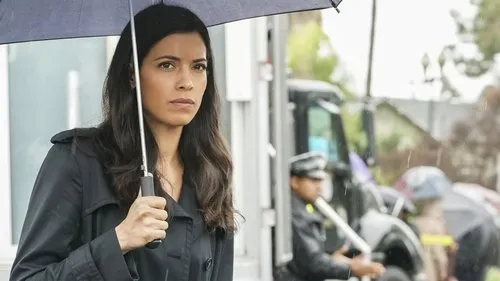 S.W.A.T.:Força de Intervenção – T01E20:Vingança 1 Mulher morena de expressão séria,em trench coat escuro,sob guarda-chuva na chuva. Fundo borrado com figura fardada e viatura.