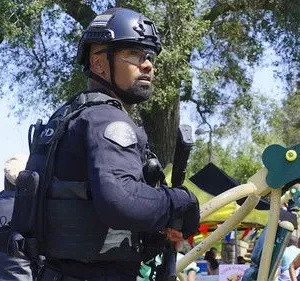 Oficial tático com capacete e óculos, em guarda, em um parque ensolarado com playground e pessoas ao fundo.