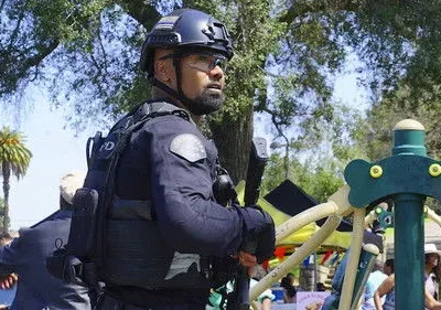 Oficial tático com capacete e óculos, em guarda, em um parque ensolarado com playground e pessoas ao fundo.