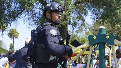 S.W.A.T.:Força de Intervenção – T01E22:Enganação 1 Oficial tático com capacete e óculos,em guarda,em um parque ensolarado com playground e pessoas ao fundo.