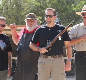 Quatro homens com armas variadas, incluindo uma espingarda e revólveres antigos, posam ao ar livre. Barbas, chapéus e óculos de sol dão um ar rústico.