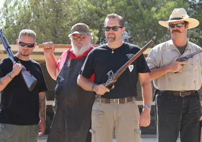 Quatro homens com armas variadas, incluindo uma espingarda e revólveres antigos, posam ao ar livre. Barbas, chapéus e óculos de sol dão um ar rústico.