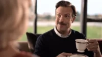 Homem de bigode, suéter azul, sorri enquanto segura uma xícara de chá. Ele olha para uma pessoa com cabelos loiros. Ambiente interno aconchegante.