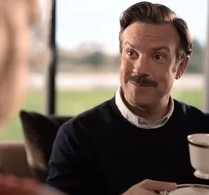 Homem de bigode, suéter azul, sorri enquanto segura uma xícara de chá. Ele olha para uma pessoa com cabelos loiros. Ambiente interno aconchegante.