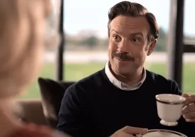 Homem de bigode, suéter azul, sorri enquanto segura uma xícara de chá. Ele olha para uma pessoa com cabelos loiros. Ambiente interno aconchegante.