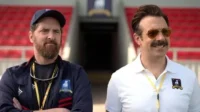 Dois homens em um estádio. Um barbudo sério de boné e um sorridente com óculos de sol e bigode, ambos com uniforme de time.