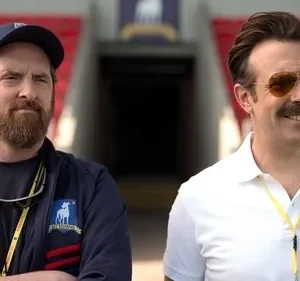 Dois homens em um estádio. Um barbudo sério de boné e um sorridente com óculos de sol e bigode, ambos com uniforme de time.