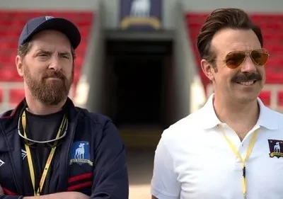 Dois homens em um estádio. Um barbudo sério de boné e um sorridente com óculos de sol e bigode, ambos com uniforme de time.