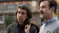 Dois homens ao ar livre. Um de cabelo grisalho longo, pensativo. O outro tem bigode, suéter e um nariz vermelho. Clima peculiar.