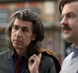 Dois homens ao ar livre. Um de cabelo grisalho longo, pensativo. O outro tem bigode, suéter e um nariz vermelho. Clima peculiar.