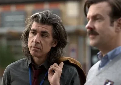 Dois homens ao ar livre. Um de cabelo grisalho longo, pensativo. O outro tem bigode, suéter e um nariz vermelho. Clima peculiar.