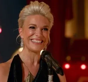 Mulher loira sorridente com penteado elegante em palco escuro com microfone. Luzes vermelhas de fundo criam clima festivo.