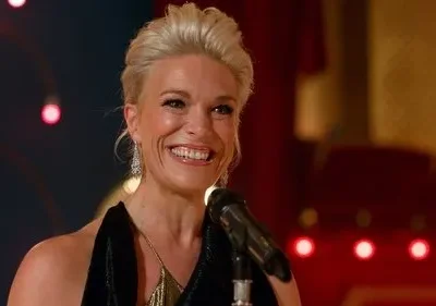 Mulher loira sorridente com penteado elegante em palco escuro com microfone. Luzes vermelhas de fundo criam clima festivo.