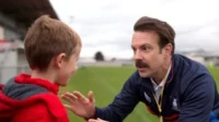 Um homem com bigode, de agasalho azul, fala com um menino de jaqueta vermelha em um campo. Ele gesticula de forma amigável.